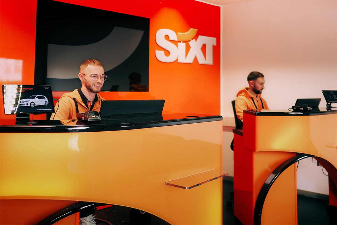 Zwei Mitarbeiter sitzen an orangefarbenen Empfangstheken vor einer orangefarbenen Wand mit dem weißen Schriftzug 'Sixt'